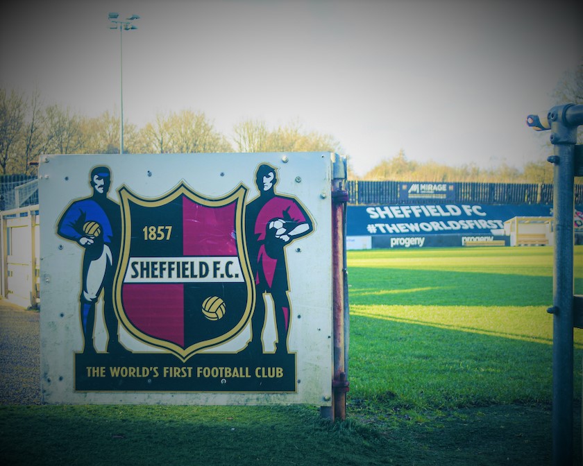 Signboard featuring the Sheffield F.C. crest with text ‘1857 Sheffield F.C.’ and ‘The World’s First Football Club,’ flanked by illustrations of two footballers. In the background, a football pitch and advertising boards display ‘Sheffield FC #TheWorldsFirst.’ Trees and sky are visible beyond the stands.