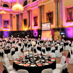 Elegant banquet hall with high ornate ceilings, chandeliers, and large portraits on the walls. Round tables are set for a formal dinner with white chair covers tied with black bows, black tablecloths, and red menus. A projector screen at the front displays “Sheffield Ambassador Dinner 2015.” Warm lighting with pink and orange uplights creates a vibrant atmosphere.