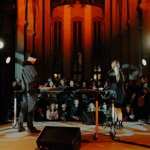 Two performers stand behind a DJ table in a grand hall with tall columns and arched windows, illuminated by warm orange lighting and spotlights. A seated audience watches in the background.