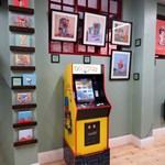 A Pac-Man arcade machine in the Pete Mckee Gallery, surrounded by prints on the wall.
