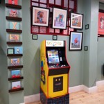 A Pac-Man arcade machine in the Pete Mckee Gallery, surrounded by prints on the wall.