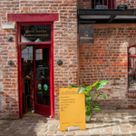 Exterior of a brick building with a red-framed door and windows. The entrance has a sign above reading “good books & other essentials,” and a hanging sign says “La Biblioteka.” A bright yellow board outside lists items such as books, magazines, stationery, prints, and gifts. A green potted plant sits beside the board on a cobbled walkway, creating a welcoming and vibrant storefront.