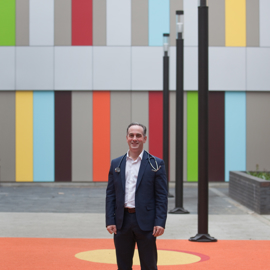 Paul Dimitri standing in a courtyard at the Sheffield Childrens Hospital. Person standing on an orange and yellow patterned outdoor surface in front of a modern building with a colorful vertical panel design in shades of green, blue, red, yellow, and gray. Several tall black lamp posts line the walkway, and the setting appears contemporary and urban.