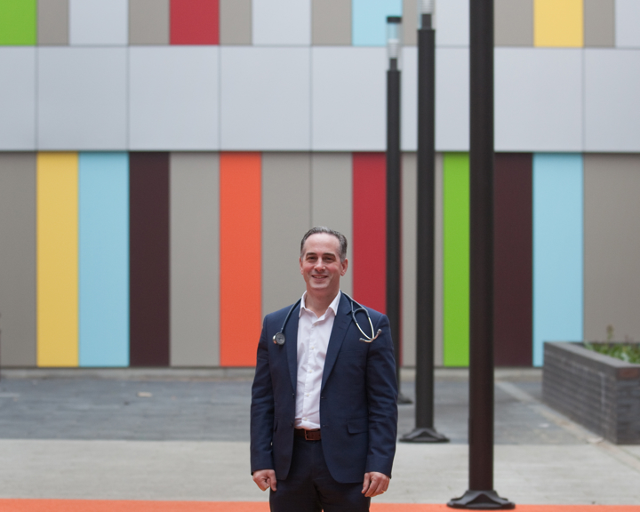 Paul Dimitri standing in a courtyard at the Sheffield Childrens Hospital. Person standing on an orange and yellow patterned outdoor surface in front of a modern building with a colorful vertical panel design in shades of green, blue, red, yellow, and gray. Several tall black lamp posts line the walkway, and the setting appears contemporary and urban.