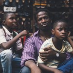 A still from the film 'My Father's Shadow', showing a man and his two sons sitting by the roadside in front of a stall.