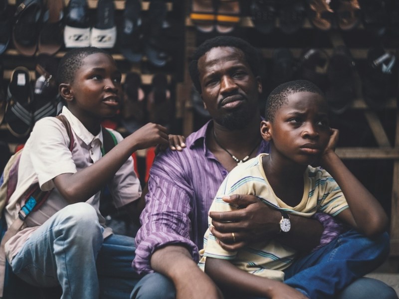 A still from the film 'My Father's Shadow', showing a man and his two sons sitting by the roadside in front of a stall.
