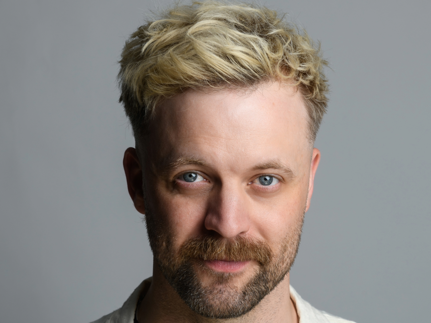 Close-up of a person with short, tousled blonde hair against a plain gray background. The individual is wearing a light-colored shirt.