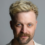 Close-up of a person with short, tousled blonde hair against a plain gray background. The individual is wearing a light-colored shirt.