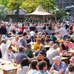 A huge crowd of people sit and stand around in a public square surrounded by street food stands on a sunny day 