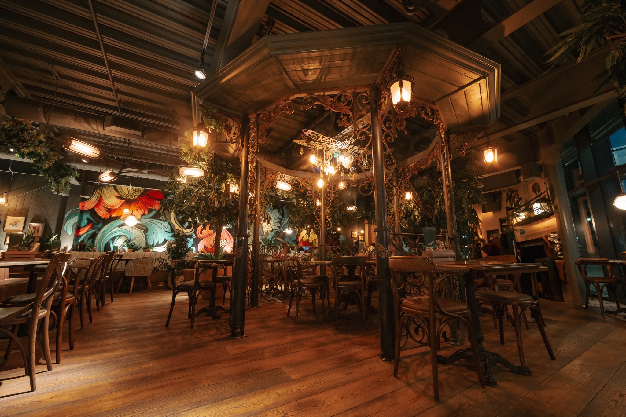 A warmly lit restaurant interior with wooden floors, wooden tables and chairs, large potted plants, and soft ambient lighting, creating a cosy, natural atmosphere.