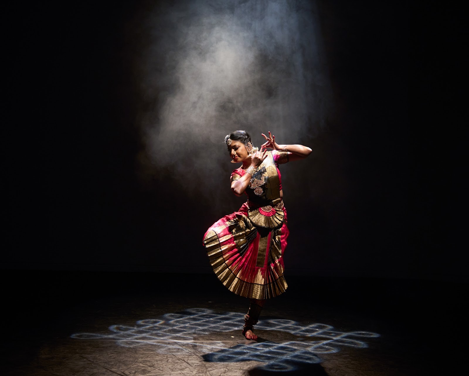 A person dances in a spotlight, in traditional Bharatanatyam dress.