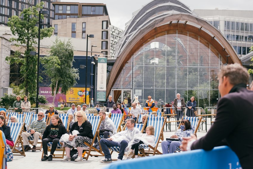 Audiences sit in deckchairs as they watch festival activity in Tudor Square, with Sheffield Winter Garden in the backdrop