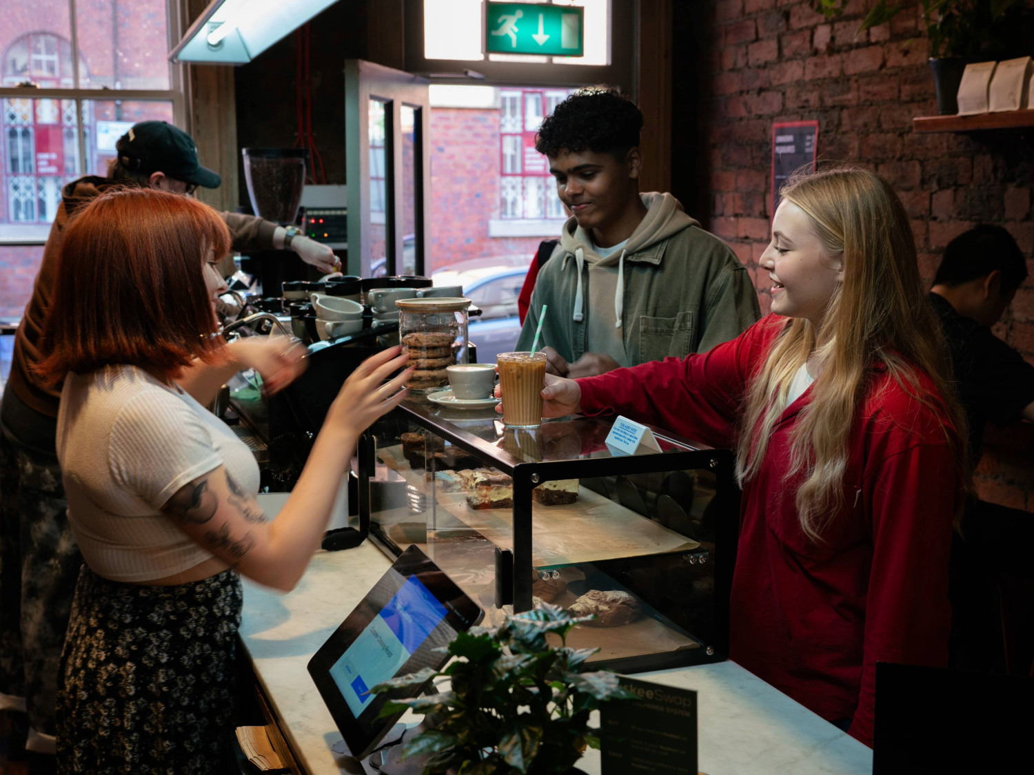 A barista is serving drinks at a coffee shop counter with a glass display case containing pastries. Two customers stand on the other side of the counter, one holding a takeaway cup with a straw. Behind the counter, coffee-making equipment and jars of biscuits are visible. The setting features exposed brick walls, a plant on the counter, and a window showing a red-brick building outside.