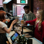 A barista is serving drinks at a coffee shop counter with a glass display case containing pastries. Two customers stand on the other side of the counter, one holding a takeaway cup with a straw. Behind the counter, coffee-making equipment and jars of biscuits are visible. The setting features exposed brick walls, a plant on the counter, and a window showing a red-brick building outside.