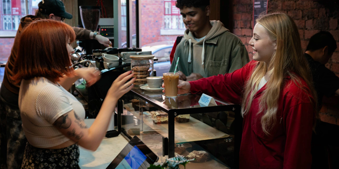 A barista is serving drinks at a coffee shop counter with a glass display case containing pastries. Two customers stand on the other side of the counter, one holding a takeaway cup with a straw. Behind the counter, coffee-making equipment and jars of biscuits are visible. The setting features exposed brick walls, a plant on the counter, and a window showing a red-brick building outside.