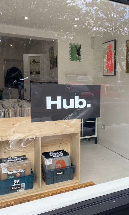 The window of a record store with a sign in the window that reads 'HUB.' . In the shop you can see wooden racks filled with vinyl records.