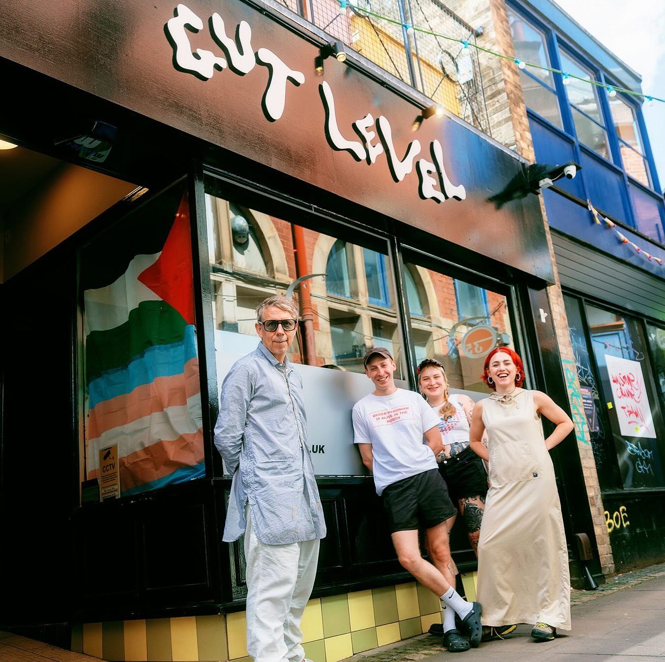 Four people standing outside a building with a black storefront sign reading ‘Gut Level’ in bold white letters. The window displays a colorful flag with horizontal stripes and a star. The group is casually dressed, and the street scene includes brick architecture and string lights above.