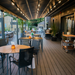 Outdoor dining area on a covered terrace with wooden flooring and black metal furniture. Round and square tables are set with small orange candle holders, creating a warm, inviting atmosphere. Overhead string lights run along the ceiling, and potted plants add greenery. The space overlooks surrounding buildings and greenery, offering a stylish and relaxed setting for alfresco dining.