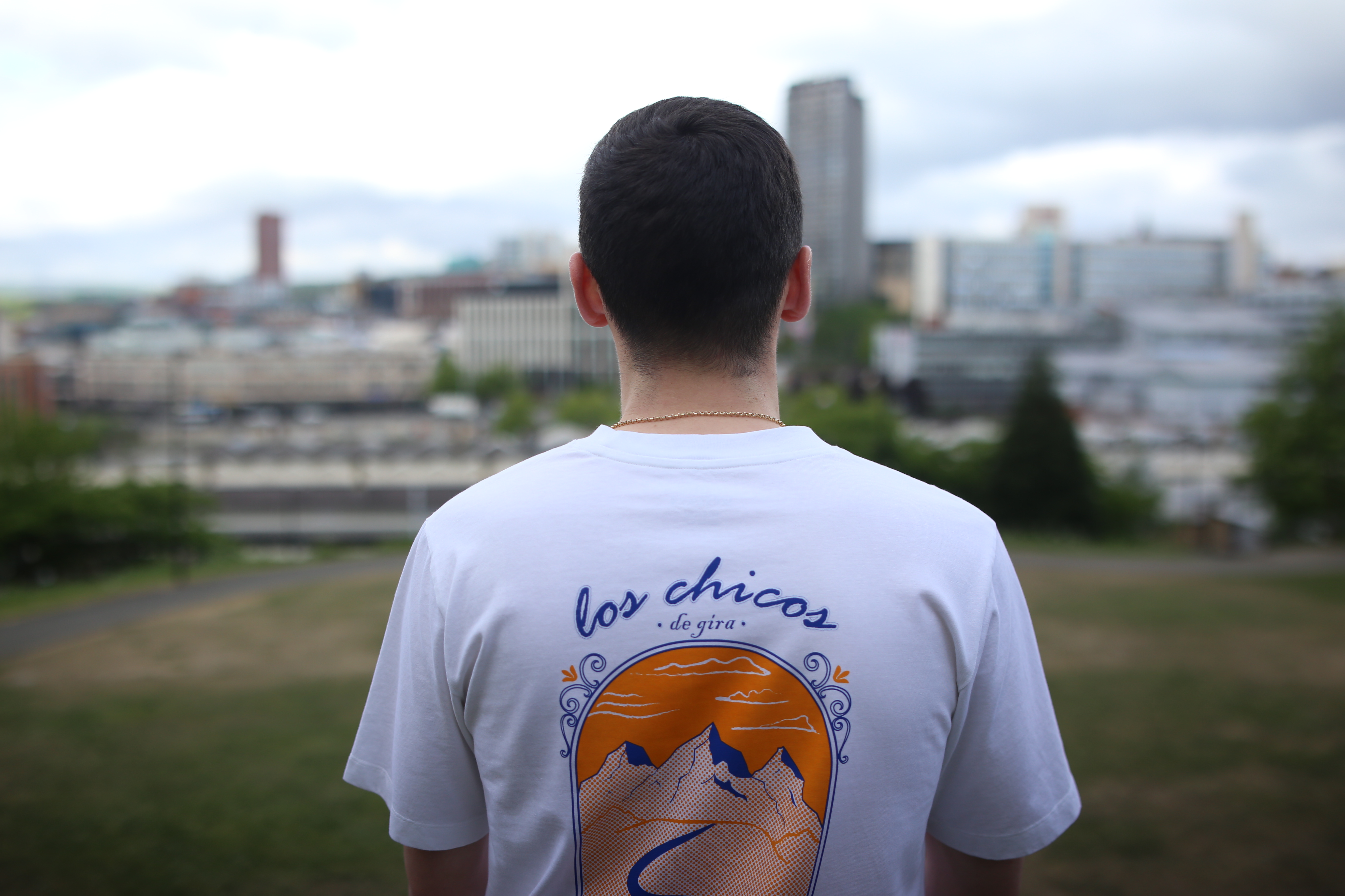  Person wearing a white T-shirt with a colorful back design showing mountains, a winding river, and an orange sunset. The text reads “los chicos de gira” in blue script. The background features a cityscape with tall buildings and greenery.