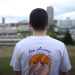  Person wearing a white T-shirt with a colorful back design showing mountains, a winding river, and an orange sunset. The text reads “los chicos de gira” in blue script. The background features a cityscape with tall buildings and greenery.