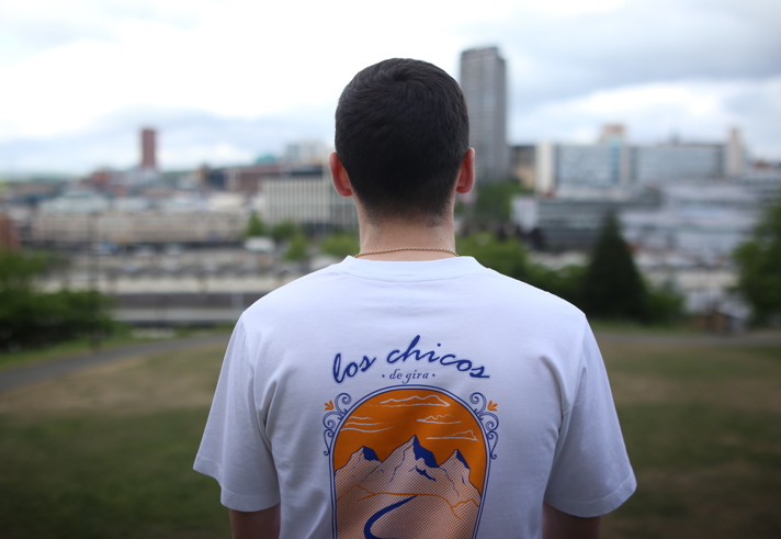  Person wearing a white T-shirt with a colorful back design showing mountains, a winding river, and an orange sunset. The text reads “los chicos de gira” in blue script. The background features a cityscape with tall buildings and greenery.