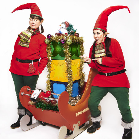 wo people dressed as festive elves in red tops, green pants, and tall red hats stand beside a decorative sleigh labeled “GOODOMETER.” The sleigh is filled with colorful gift boxes wrapped in tinsel and topped with a snowman figure. The scene is bright and cheerful, with a white background emphasising the holiday theme.