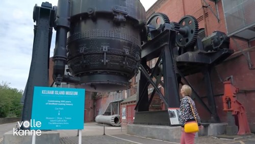 Large industrial machinery displayed outdoors at Kelham Island Museum, including a massive black steel vessel and gears mounted on a red brick building. A turquoise sign in front reads ‘Kelham Island Museum – Celebrating 300 years of Sheffield making history.’