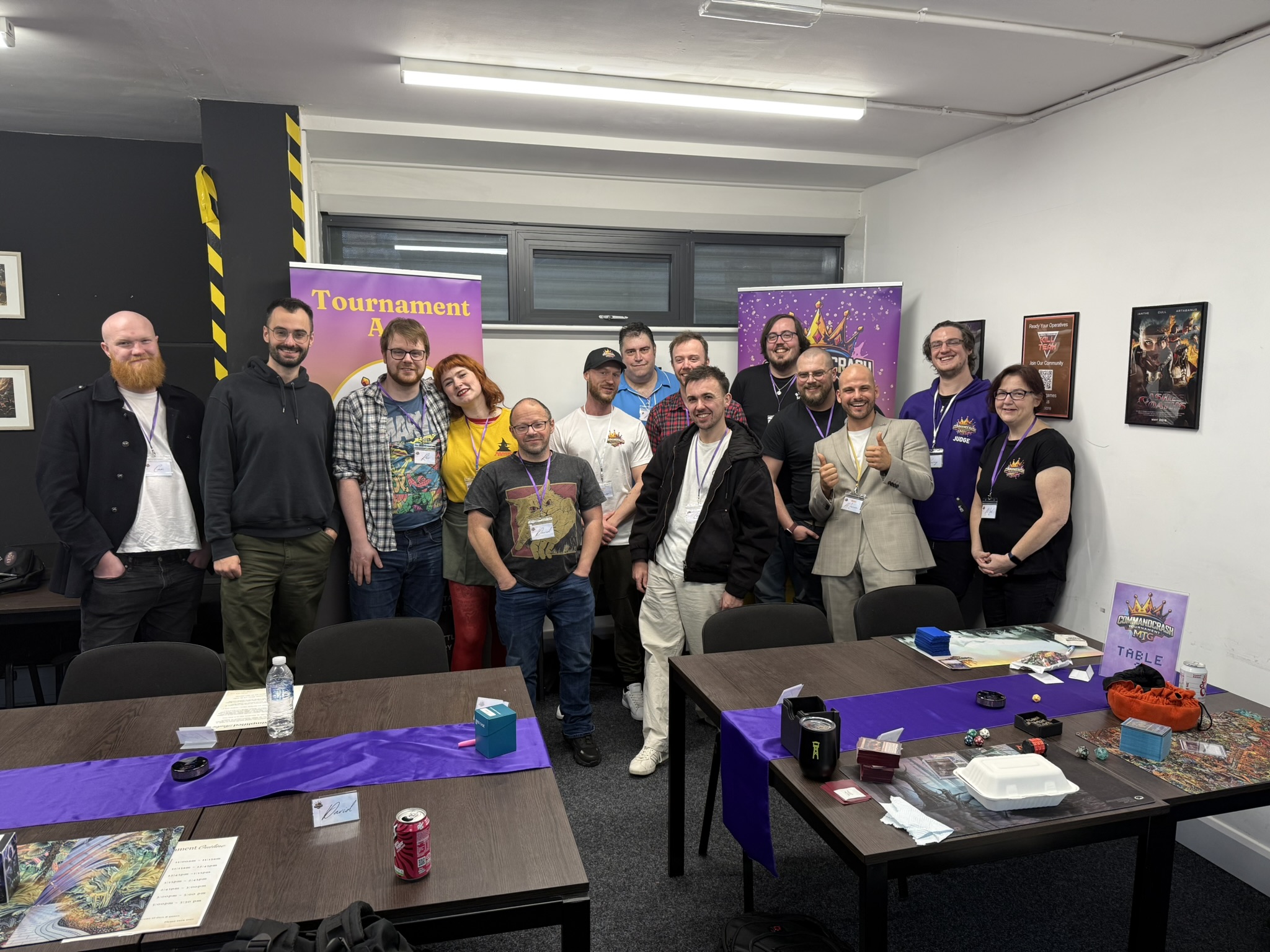 A group of people standing together in a room with tables set up for a card game event. The tables have purple table runners, cards, dice, and drinks. Behind the group are two colourful banners, one reading “Tournament Area” and the other featuring the “Command Crash MTG” logo. Framed artwork is visible on the walls, and the setting appears to be an organised gaming tournament.
