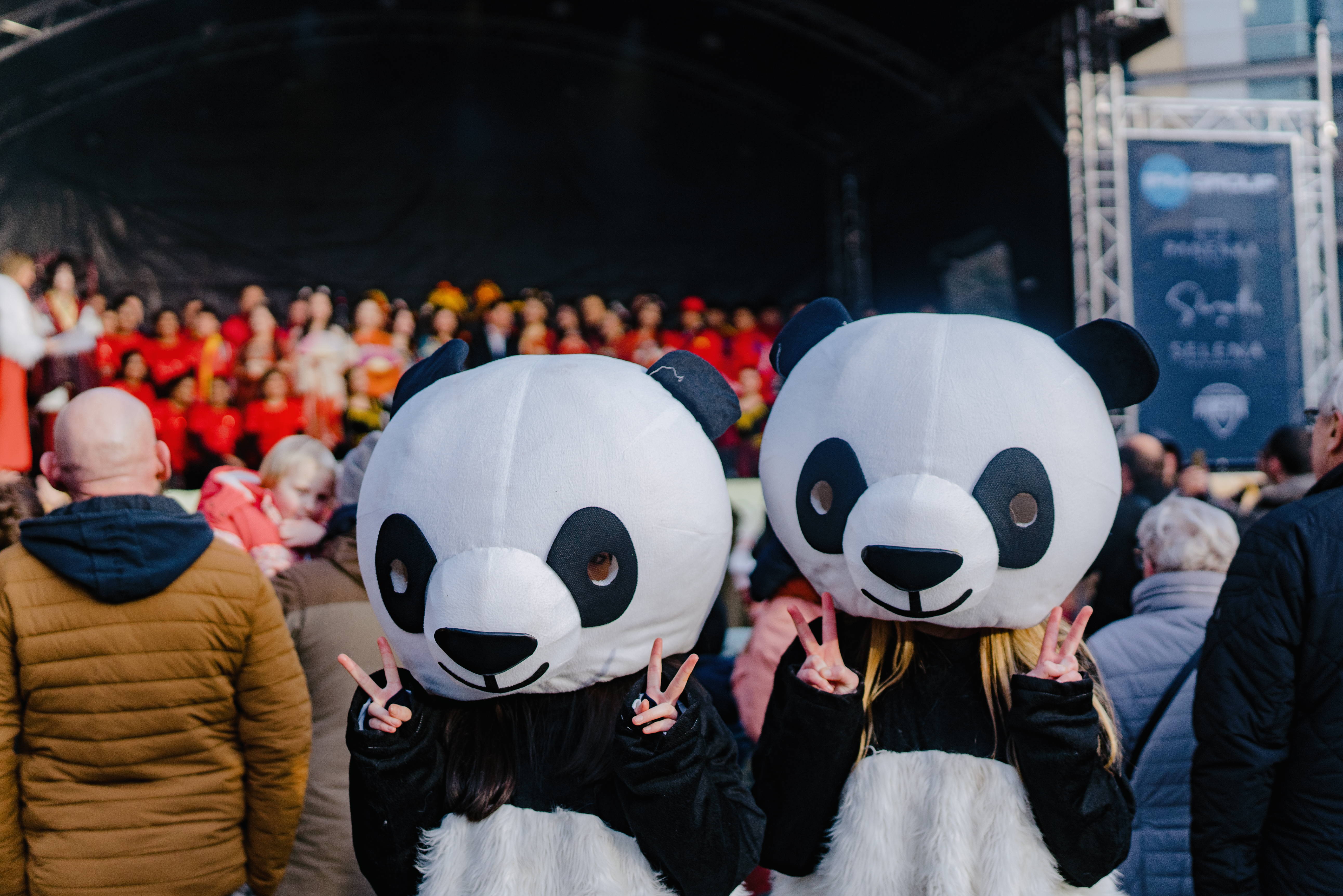  Two people wearing oversized panda costumes facing the camera, making peace signs with their hands. Behind them, a crowd watches a performance on a stage filled with participants in bright red outfits.