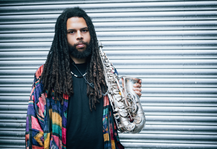 A person with long dreadlocks stands in front of a corrugated metal wall, wearing a colourful patterned jacket over a dark shirt. They hold a silver alto saxophone in one hand, angled slightly forward, with its keys and curved bell clearly visible.