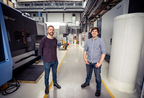 Two people standing in a spacious industrial workshop with large machinery on both sides. The environment features high ceilings, overhead beams, and bright lighting. The floor has marked yellow lines for pathways, and various equipment and workstations are visible in the background.