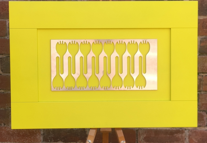Contemporary artwork displayed on an easel, featuring a bright yellow frame and a central metallic panel with cut-out fork-like shapes, set against a brick wall background.