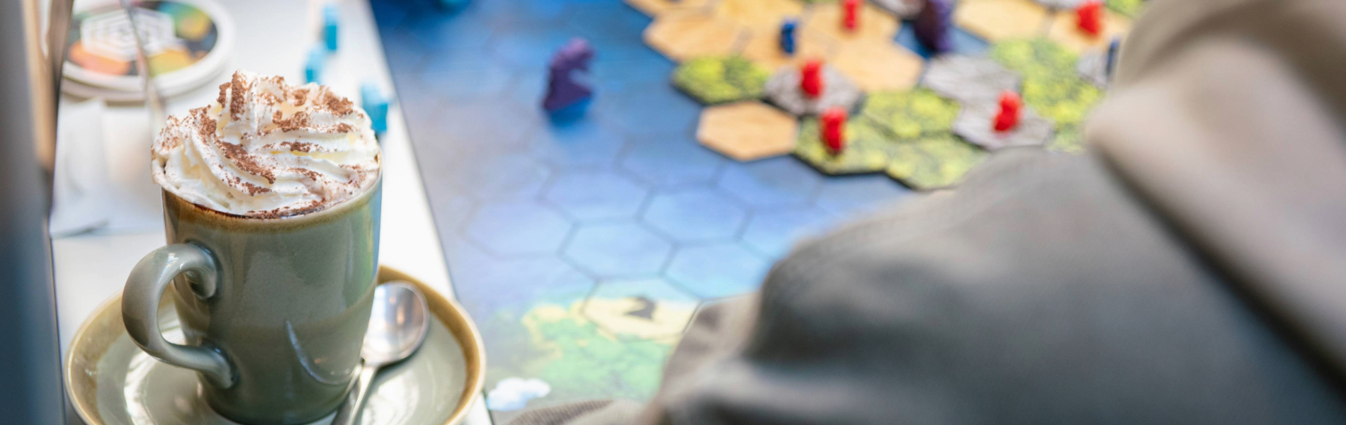 A close-up of a cappuccino sitting on a table, next to a board game. A hand is moving one of the pieces from the game. 