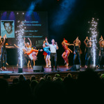 A large ensemble of performers fills a brightly lit theatre stage, dancing in formation with pyrotechnic-style spark effects rising on both sides of the stage. A person stands at the front center leading the performance. Behind them, a screen displays text and a black‑and‑white portrait. Audience silhouettes are visible at the bottom of the image Credit: Green Vision Photography