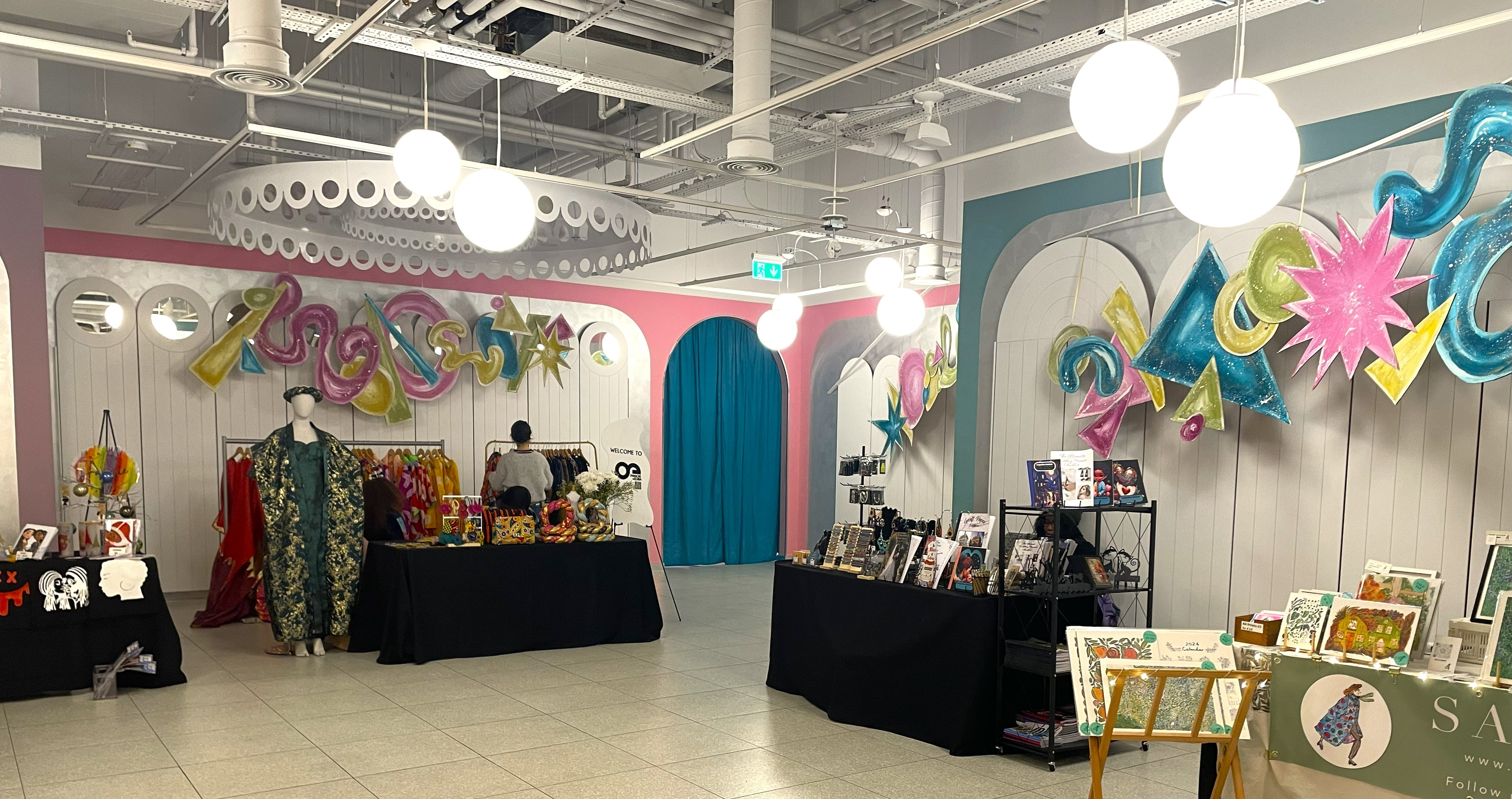 Wide view of a pop-up market interior with tables covered in black cloth displaying handmade crafts, artwork, and clothing. The space features vibrant wall decorations in pink, teal, and yellow with large hanging lights.