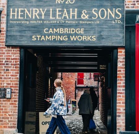Closer view of the entrance to Leah’s Yard, showing the large wooden sign for ‘Henry Leah & Sons Ltd – Cambridge Stamping Works’ above the covered passageway. A cobbled walkway continues through to the courtyard, with brick façades and red-trimmed windows visible in the background.