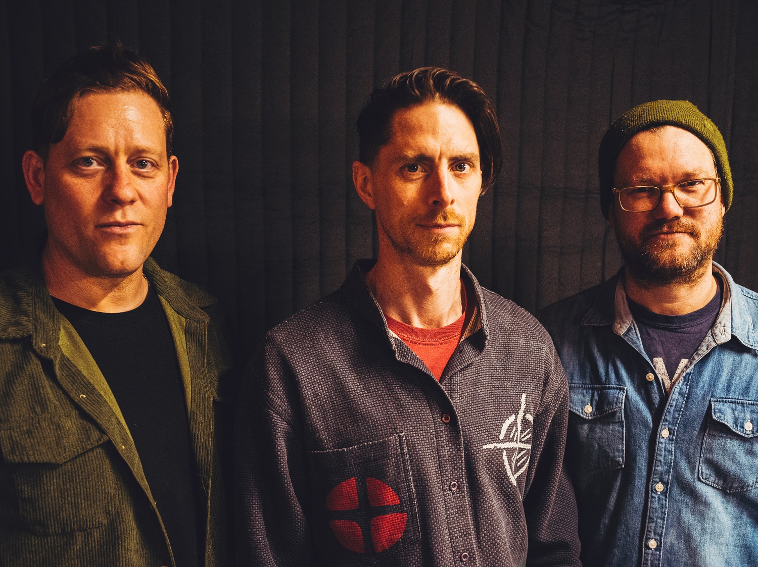 Three people standing side by side against a dark curtain backdrop. The person on the left wears an olive-green corduroy-style jacket over a dark top. The person in the middle wears a patterned shirt with a red patch design and a red T‑shirt underneath. The person on the right wears a denim shirt layered over another top and a knitted beanie.