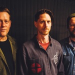 Three people standing side by side against a dark curtain backdrop. The person on the left wears an olive-green corduroy-style jacket over a dark top. The person in the middle wears a patterned shirt with a red patch design and a red T‑shirt underneath. The person on the right wears a denim shirt layered over another top and a knitted beanie.
