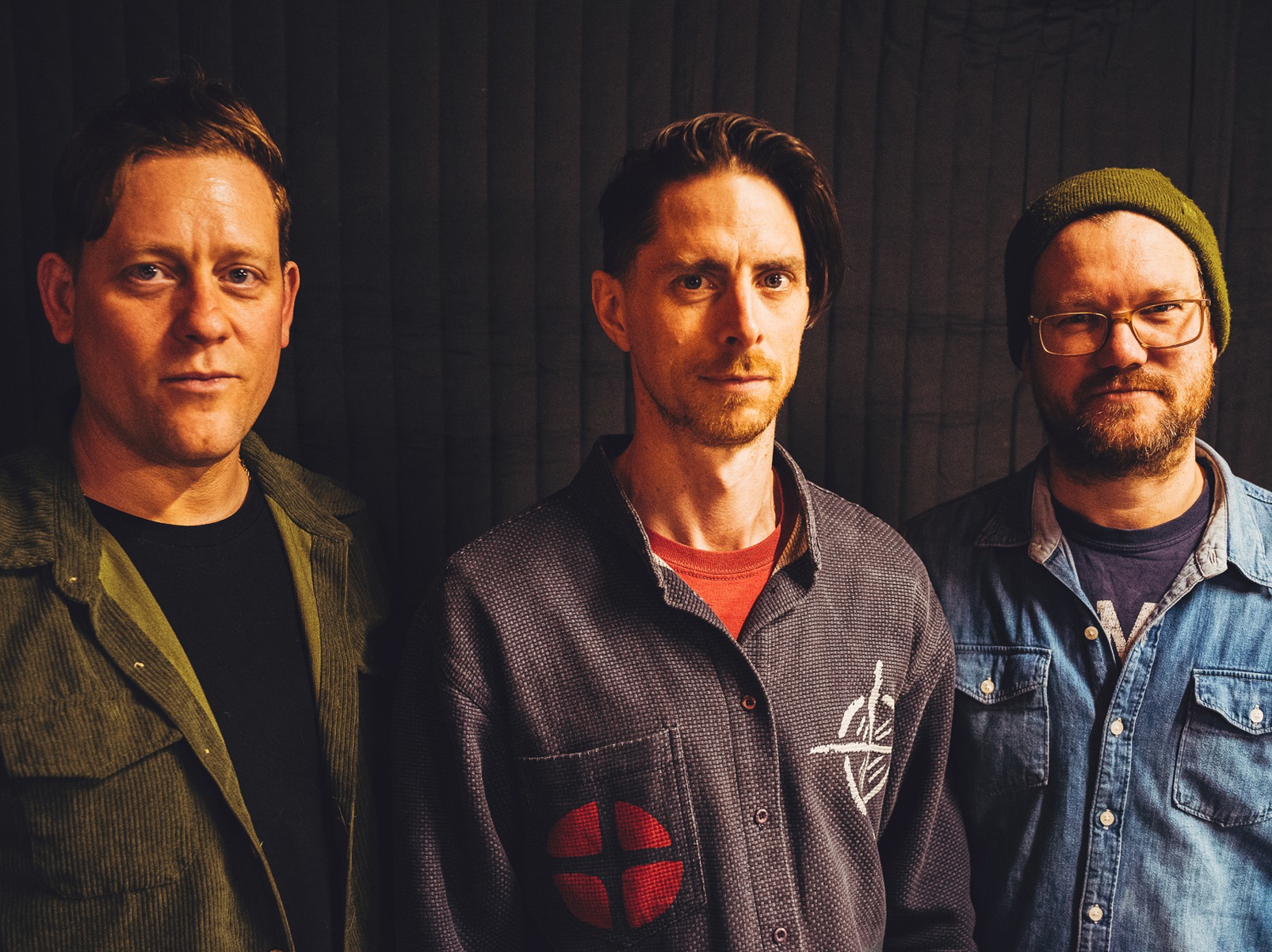 Three people standing side by side against a dark curtain backdrop. The person on the left wears an olive-green corduroy-style jacket over a dark top. The person in the middle wears a patterned shirt with a red patch design and a red T‑shirt underneath. The person on the right wears a denim shirt layered over another top and a knitted beanie.