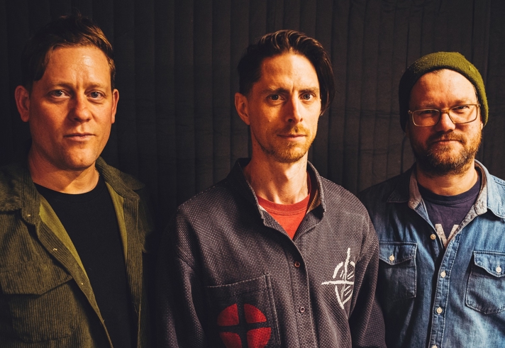Three people standing side by side against a dark curtain backdrop. The person on the left wears an olive-green corduroy-style jacket over a dark top. The person in the middle wears a patterned shirt with a red patch design and a red T‑shirt underneath. The person on the right wears a denim shirt layered over another top and a knitted beanie.