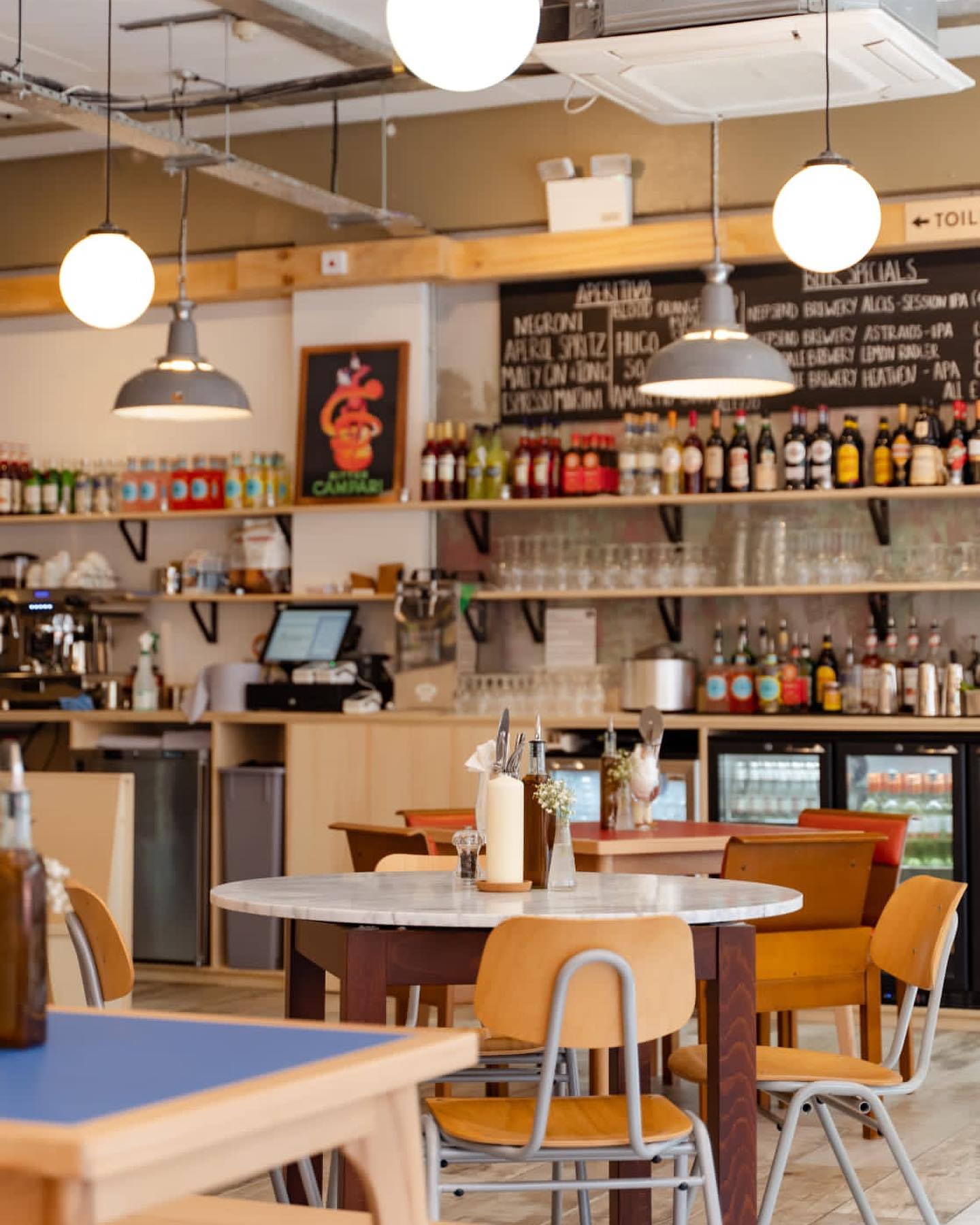 A bright, modern pizzeria interior with round tables, wooden chairs, hanging globe lights and shelves stocked with drinks behind the counter.
