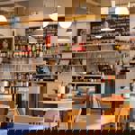A bright, modern pizzeria interior with round tables, wooden chairs, hanging globe lights and shelves stocked with drinks behind the counter.