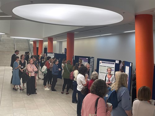A group of people are gathered in a brightly lit hallway with large circular ceiling lights and orange columns. They are viewing and discussing research posters displayed on dark blue boards arranged along the wall. The posters contain text and images, and attendees appear to be engaged in conversation, some holding drinks. A staircase is visible in the background, leading to an upper level.