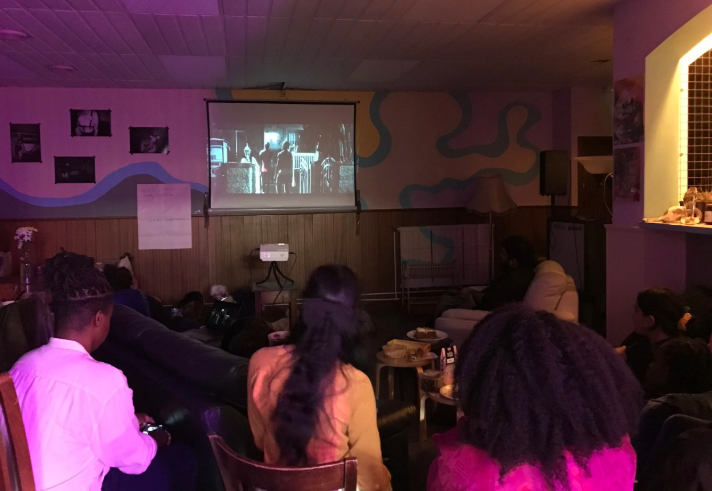 A darkened room where a film is being screened. There are rows of people sat in semi-circles, watching the screen.