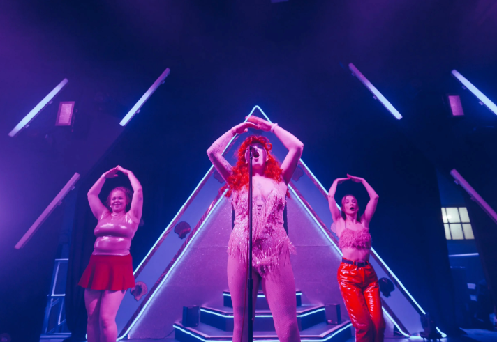 A vibrant stage performance featuring three costumed performers under purple and blue lighting. The central performer stands in front of a microphone wearing a fringed bodysuit and a bright red wig, framed by a large illuminated triangular backdrop. Two other performers stand on either side, dressed in shiny outfits, with arms raised above their heads in a matching pose. Neon light bars and stage equipment accentuate the dramatic, colourful setting.