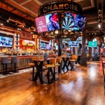 A spacious, modern bar interior with wooden floors, high tables, and cushioned stools. Multiple large screens display sports and live band promotions. The central feature is a circular structure above the bar with a large dartboard graphic. Warm lighting and industrial-style décor create an energetic atmosphere.