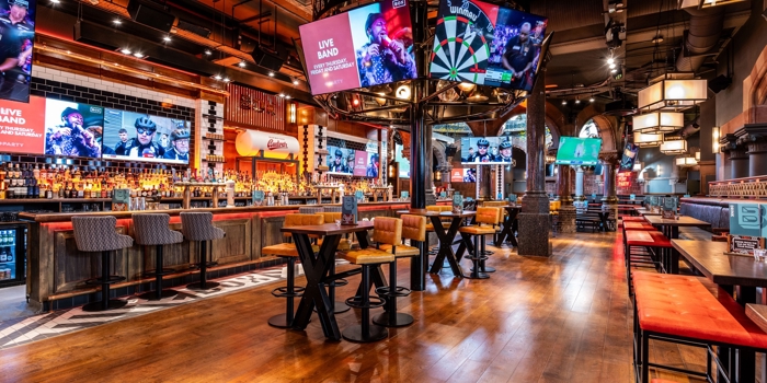 A spacious, modern bar interior with wooden floors, high tables, and cushioned stools. Multiple large screens display sports and live band promotions. The central feature is a circular structure above the bar with a large dartboard graphic. Warm lighting and industrial-style décor create an energetic atmosphere.