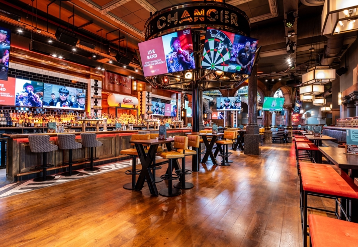 A spacious, modern bar interior with wooden floors, high tables, and cushioned stools. Multiple large screens display sports and live band promotions. The central feature is a circular structure above the bar with a large dartboard graphic. Warm lighting and industrial-style décor create an energetic atmosphere.