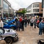 The custom car show from a previous year, with crowds of people looking around cars on display.