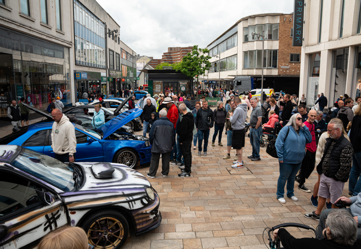 The custom car show from a previous year, with crowds of people looking around cars on display.